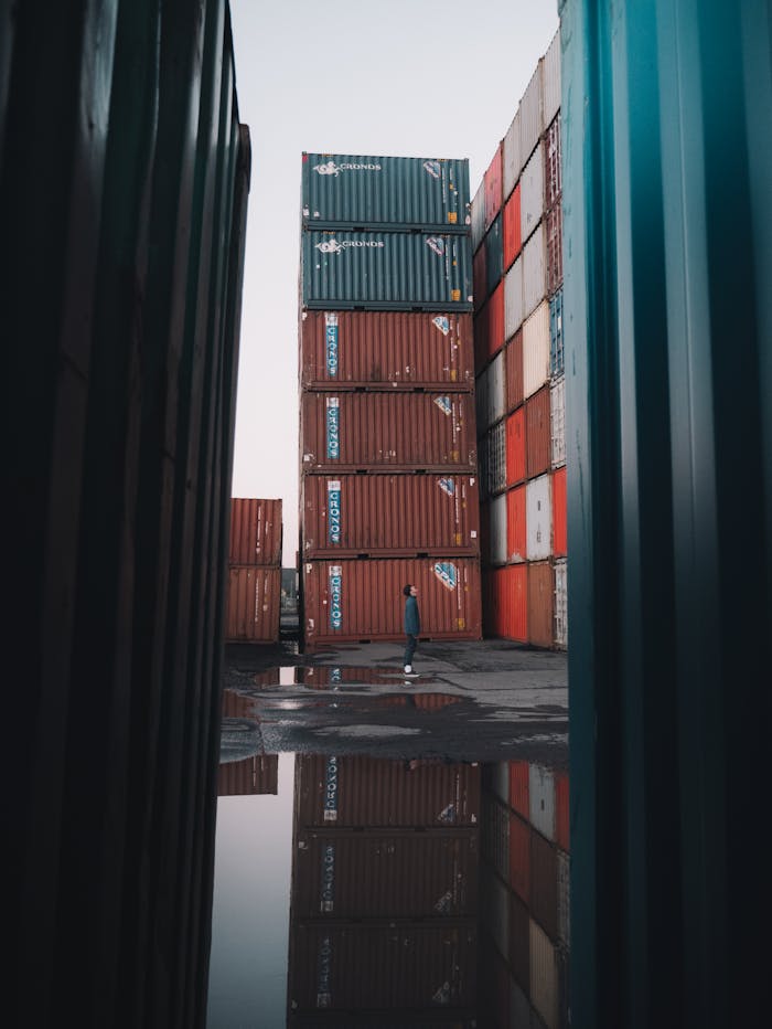 A solitary person walks between stacked shipping containers with reflections in water.
